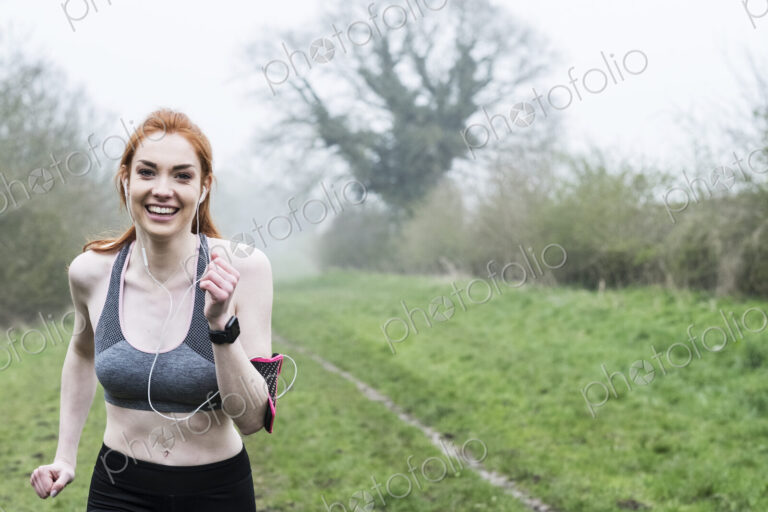 Smiling young woman with long red hair wearing sportswear, exercising outdoors,…