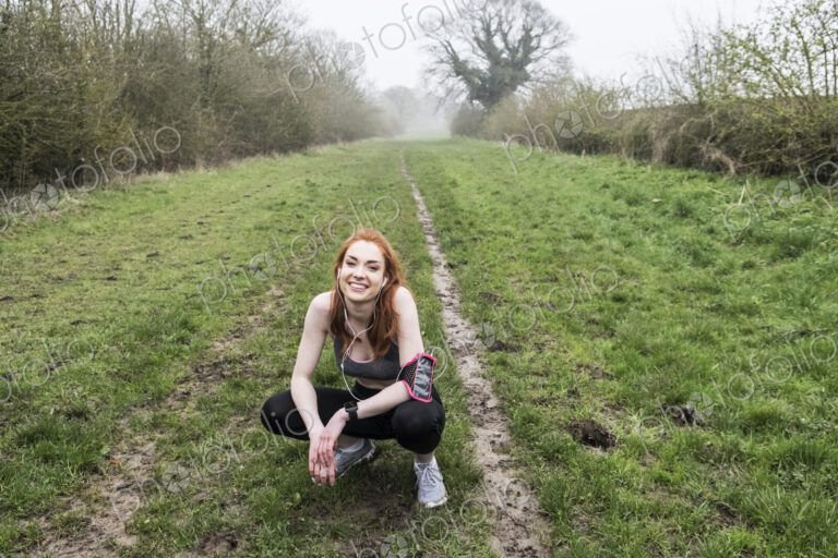 Smiling young woman with long red hair wearing sportswear, exercising outdoors,…