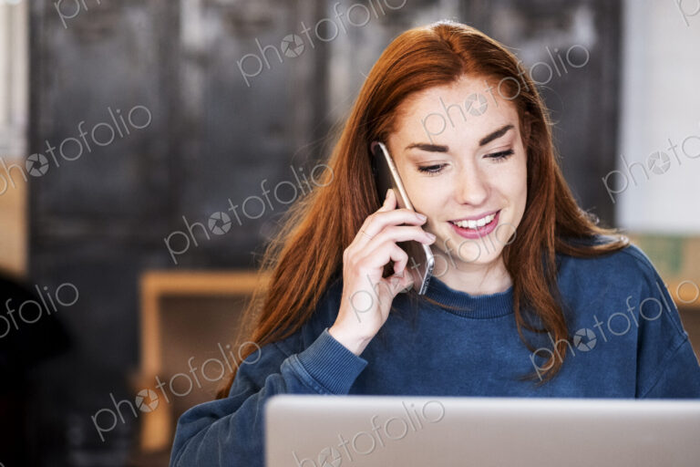 Smiling young woman working on laptop using mobile phone.