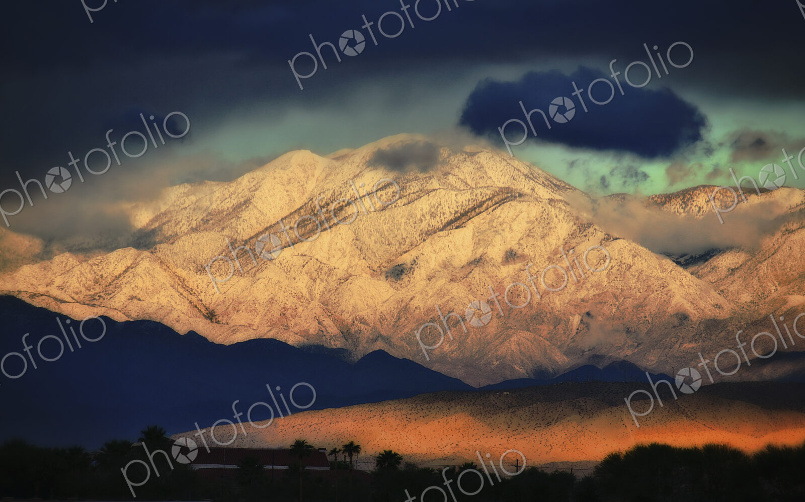 Snowcovered slopes of a mountain range with a dark stormy sky above, and a low sunlit…