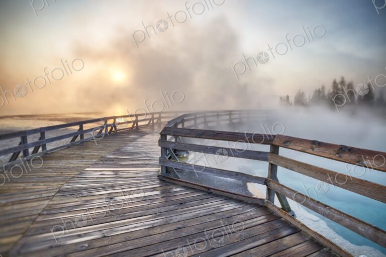 Steaming hot spring at sunrise with wooden boardwalk curving through West Thumb Geyser…