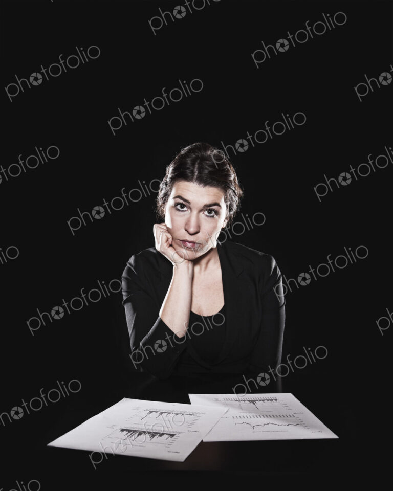 Studio portrait, a woman sitting with paperwork spread out and her hand on her chin,…