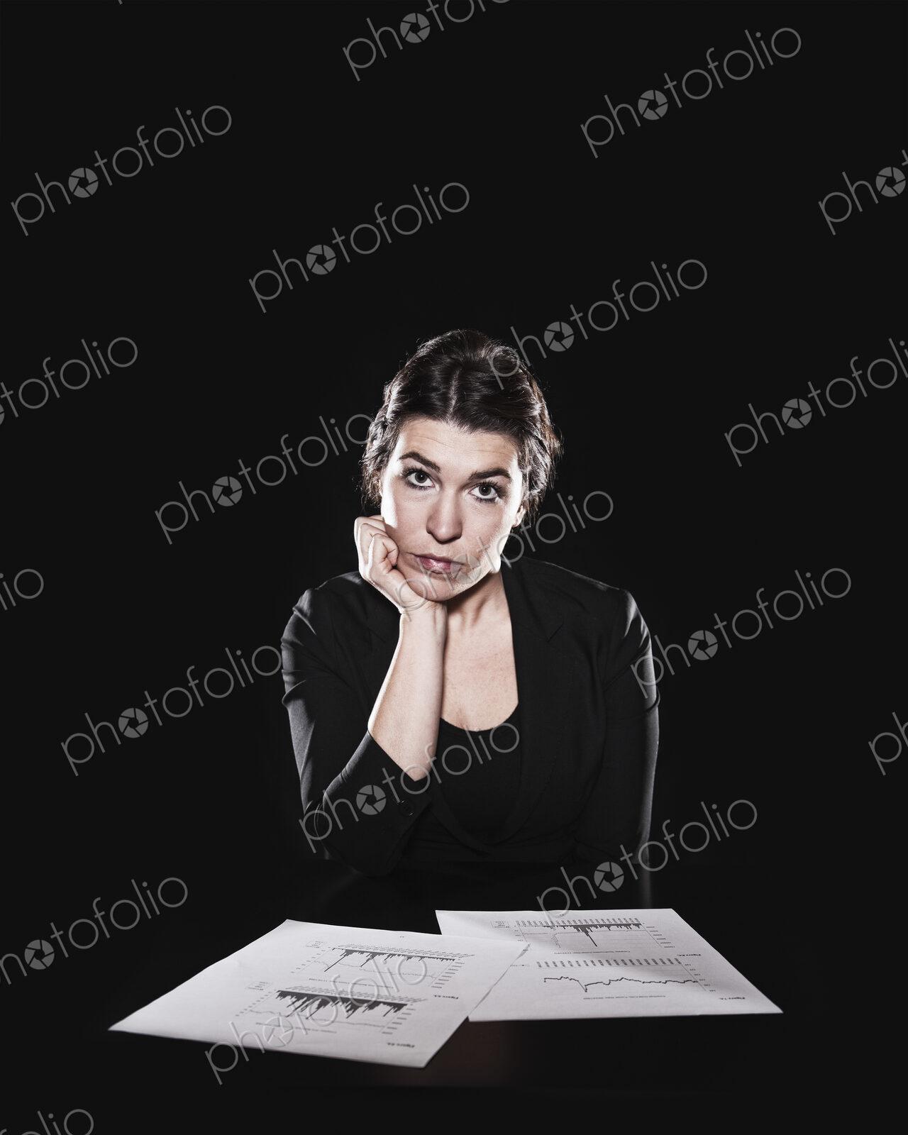Studio portrait, a woman sitting with paperwork spread out and her hand on her chin,…