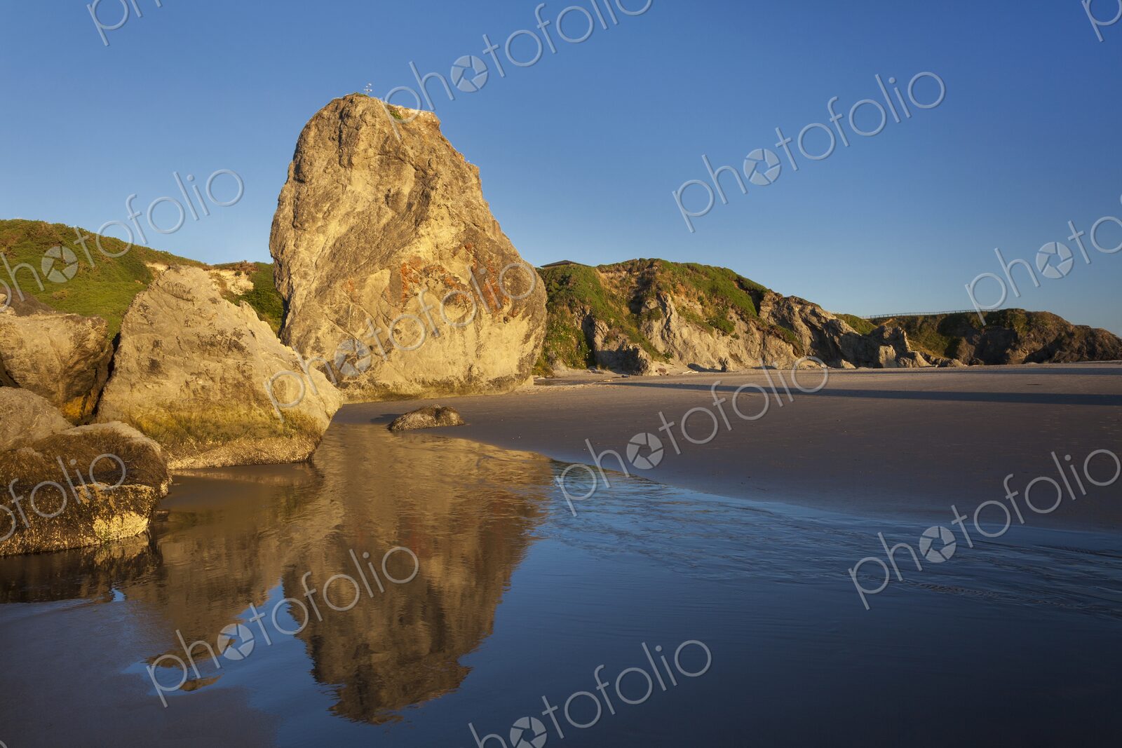 Sunlit rock formations reflect in the wet sand at low tide along the Oregon Coast in…