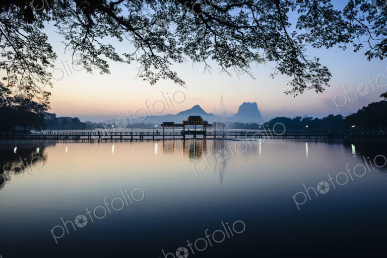 Sunrise across a calm lake, a footbridge and pavilion, mountains in the distance.