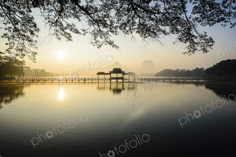 Sunrise across a calm lake, a footbridge and pavilion, mountains in the distance.