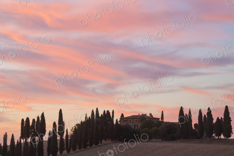 Sunset across a cloudy Italian landscape with cypress trees and farmhouse.