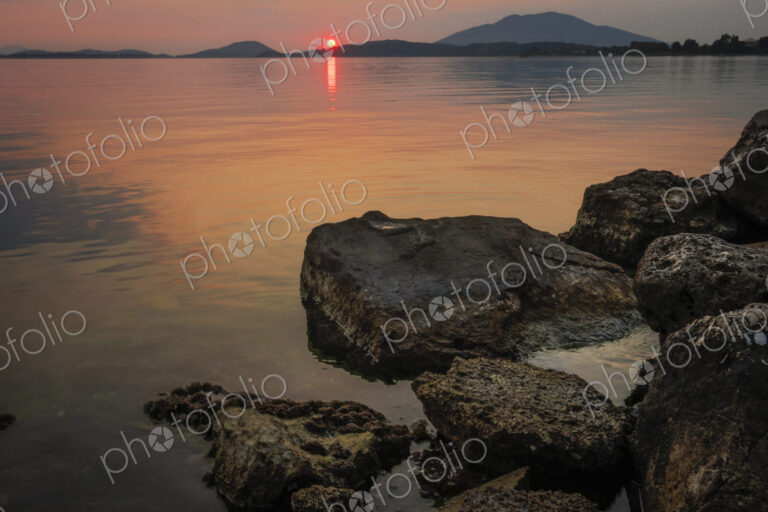 Sunset over a flat calm sea and view of land masses on the horizon.