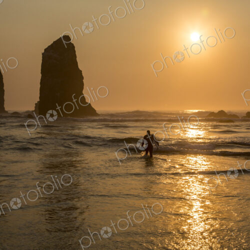 Surfer walking out of the sea with sun setting behind rock in waves on beach.