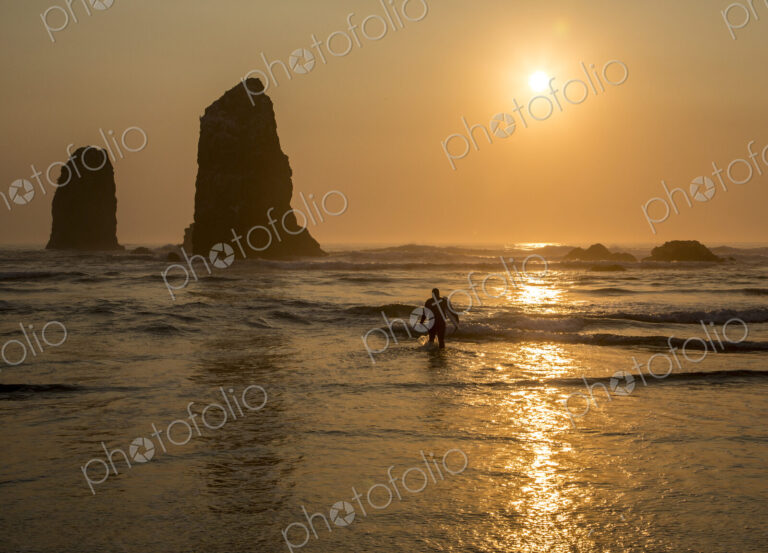 Surfer walking out of the sea with sun setting behind rock in waves on beach.