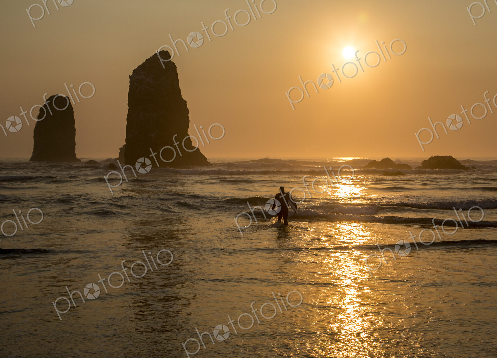 Surfer walking out of the sea with sun setting behind rock in waves on beach.