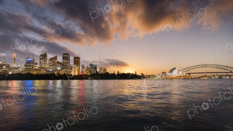 Sydney lit up at dawn seen from the water, including the Sydney Opera House.