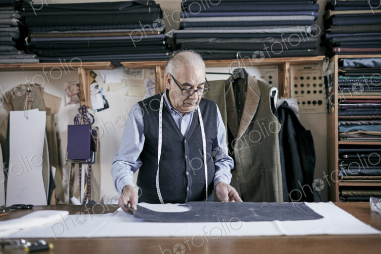 Tailor with measuring tape around neck working on a cut piece of fabric.