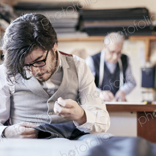 Tailor with measuring tape around neck working with material at bench Tailor with measuring tape around neck working with material at bench