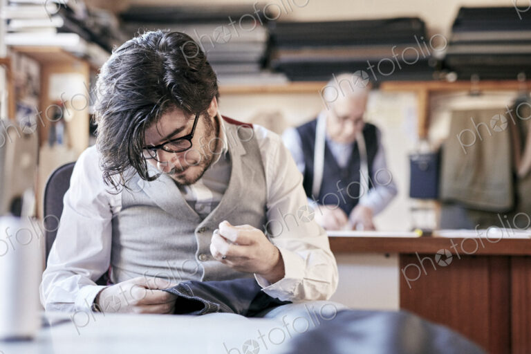 Tailor with measuring tape around neck working with material at bench