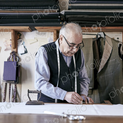 Tailor with measuring tape around neck working with material at bench Tailor with measuring tape around neck working with material at bench