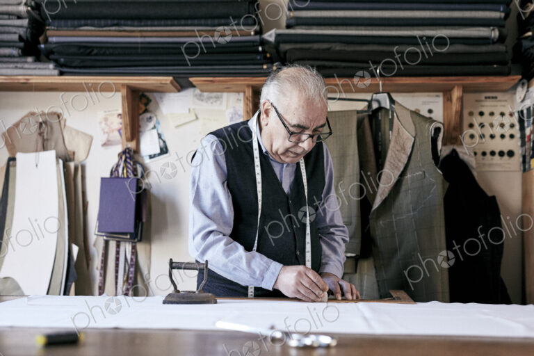 Tailor with measuring tape around neck working with material at bench