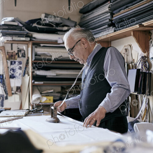 Tailor with measuring tape around neck working with material at bench Tailor with measuring tape around neck working with material at bench