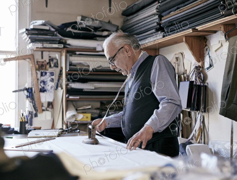 Tailor with measuring tape around neck working with material at bench