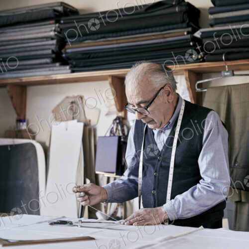 Tailor with measuring tape around neck working with material at bench Tailor with measuring tape around neck working with material at bench