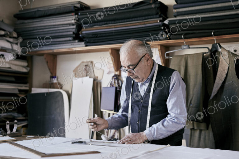 Tailor with measuring tape around neck working with material at bench