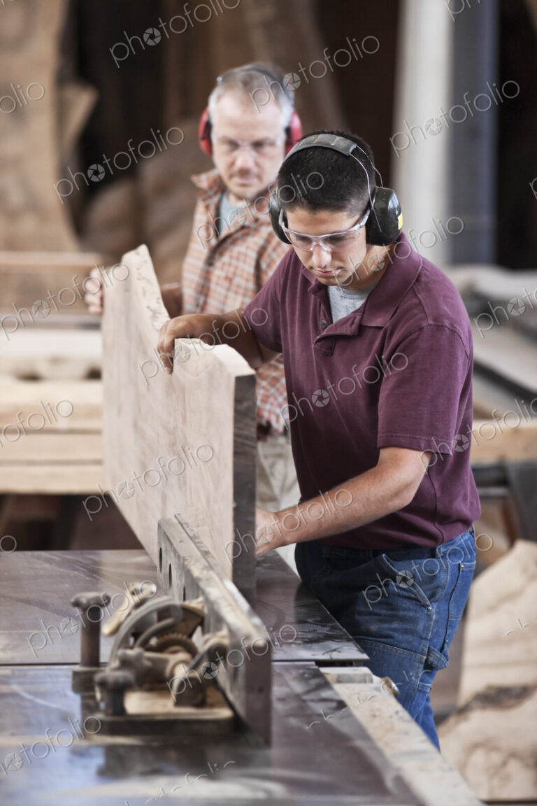 Team of two caucasian men cutting a large wooden slab on a table saw in a woodworking…