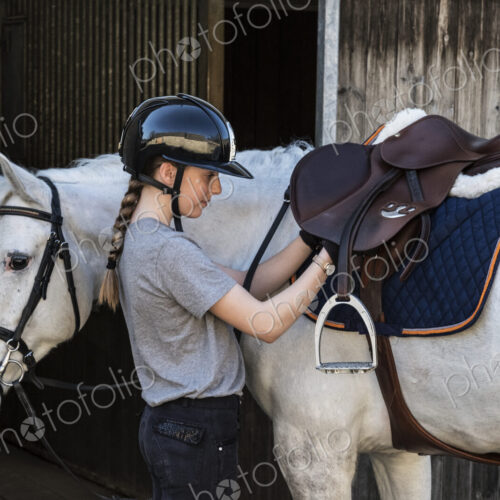Teenage girl horserider with a grey horse outside astable, adjusting the girth and saddle.