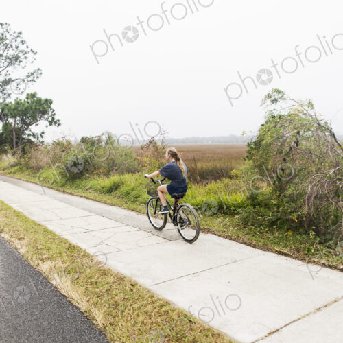Teenage girl riding a bicycle along a path on open ground by water, St. Simon's… Teenage girl riding a bicycle along a path on open ground by water, St. Simon's…