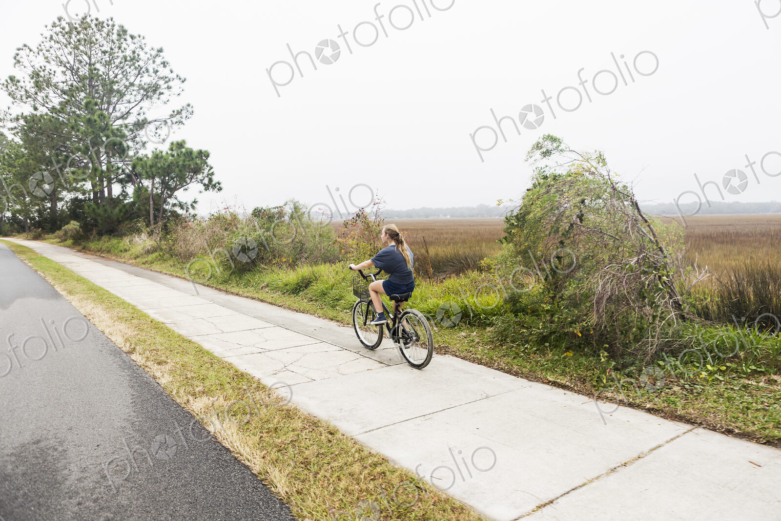 Teenage girl riding a bicycle along a path on open ground by water, St. Simon’s…