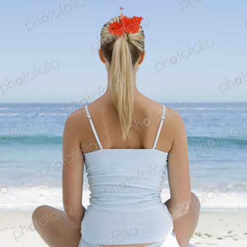 The beach. A young woman seated on a towel on the sand in a relaxed pose, looking out…