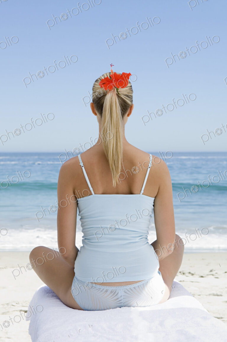 The beach. A young woman seated on a towel on the sand in a relaxed pose, looking out…