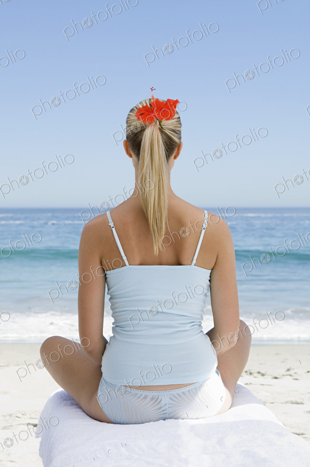 The beach. A young woman seated on a towel on the sand in a relaxed pose, looking out…