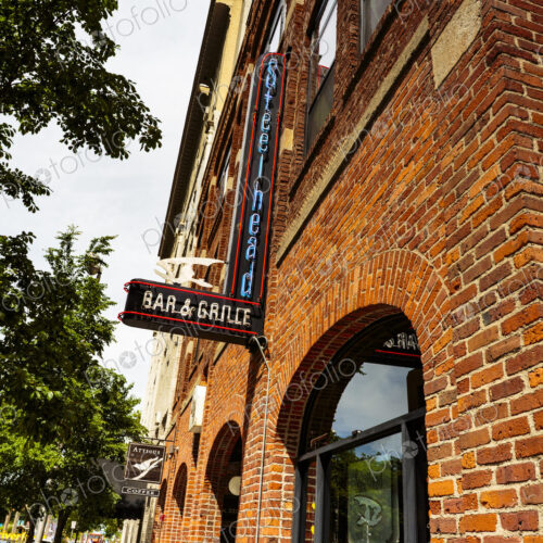 The exterior of a bar, a red brick building, neon signs, and an A board advertising…