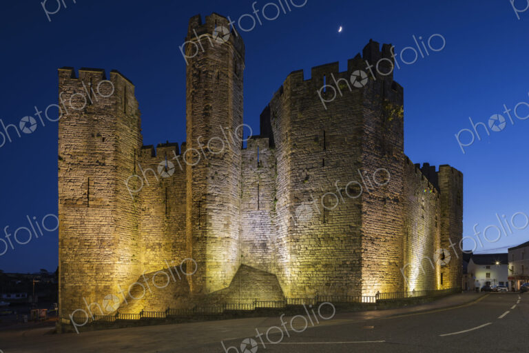 The historic Norman motte and bailey Caernarfon Castle, walls lit up at dusk.