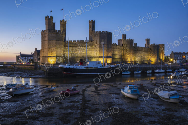 The historic Norman motte and bailey Caernarfon Castle, walls lit up at dusk, the…
