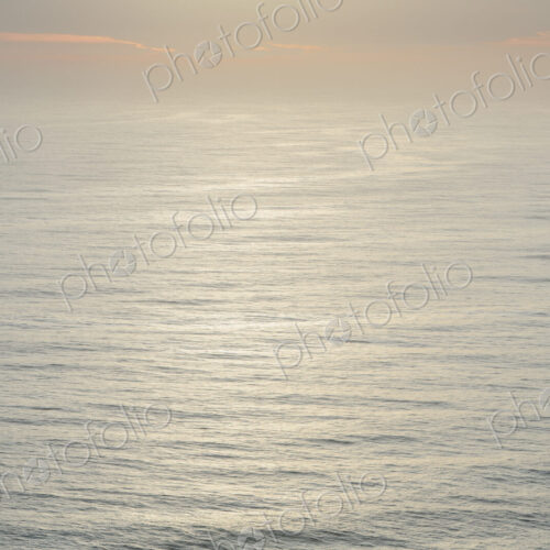 The open ocean, water surface calm and grey and the glow of the sun at dawn on the…