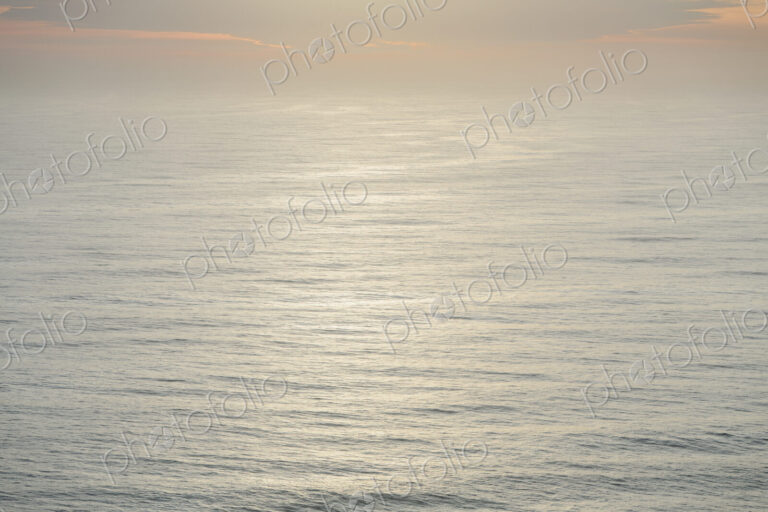 The open ocean, water surface calm and grey and the glow of the sun at dawn on the…