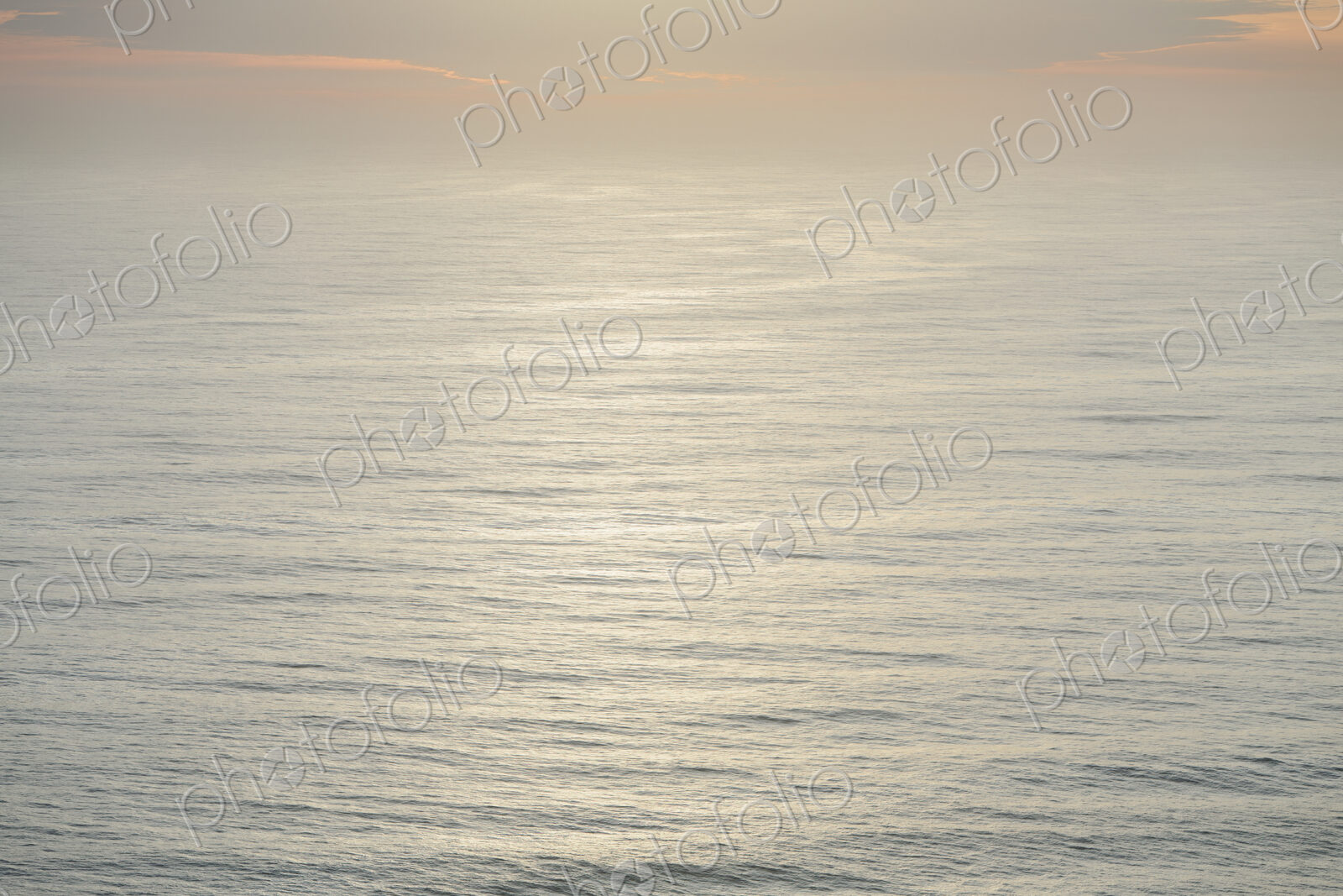 The open ocean, water surface calm and grey and the glow of the sun at dawn on the…
