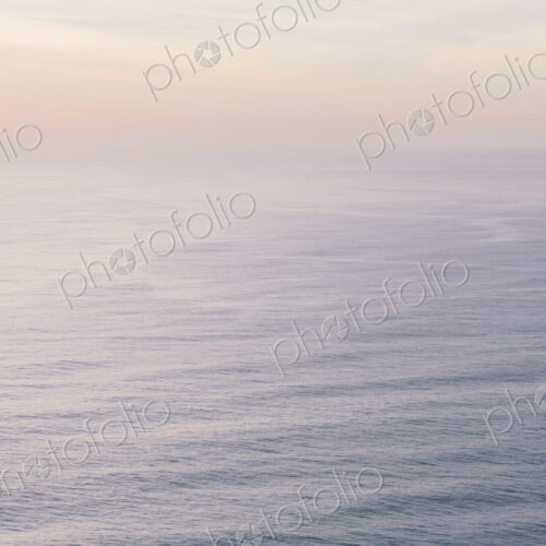 The open ocean, water surface calm and grey and the glow of the sun at dawn on the…