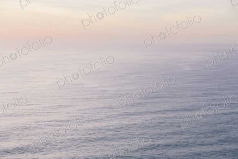 The open ocean, water surface calm and grey and the glow of the sun at dawn on the…
