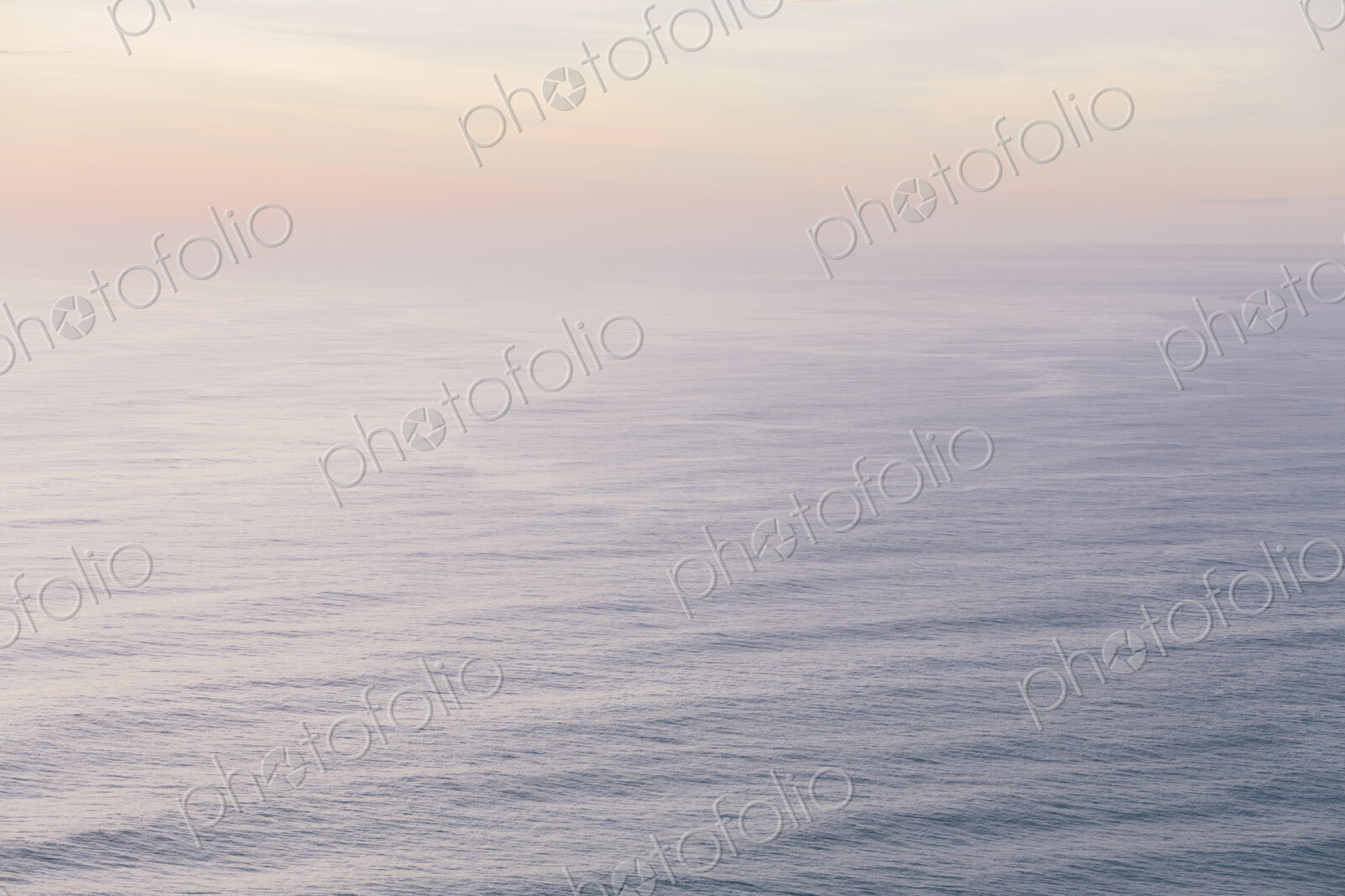 The open ocean, water surface calm and grey and the glow of the sun at dawn on the…