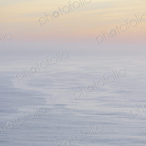 The open ocean, water surface calm and grey and the glow of the sun at dawn on the…