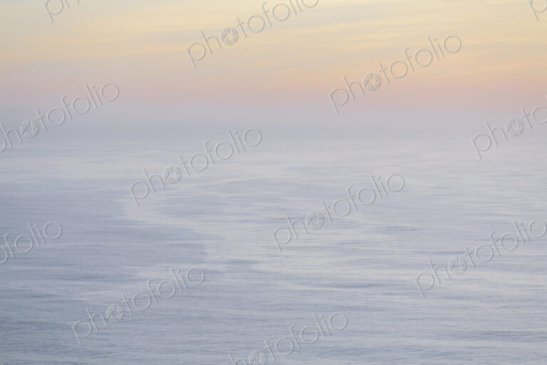 The open ocean, water surface calm and grey and the glow of the sun at dawn on the…