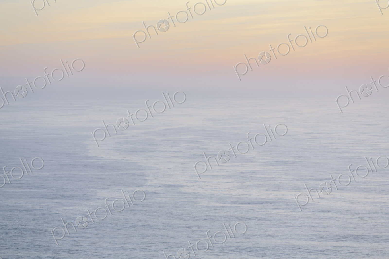 The open ocean, water surface calm and grey and the glow of the sun at dawn on the…