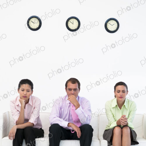 Three business people sitting on couch under clocks looking nervous.