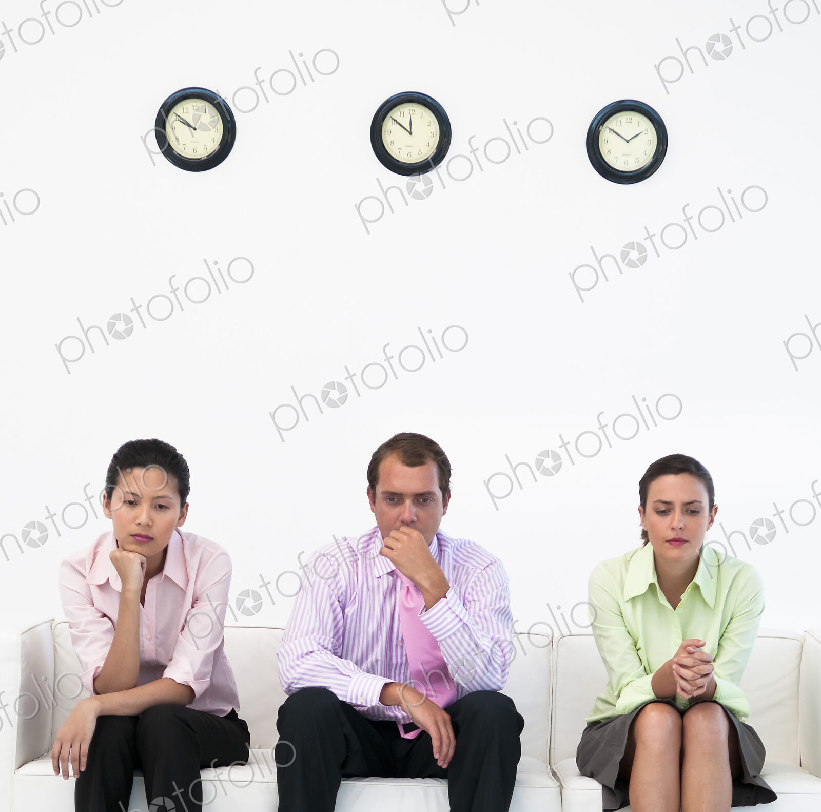 Three business people sitting on couch under clocks looking nervous.
