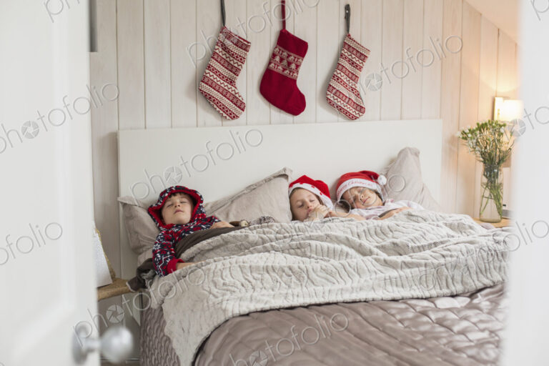 Three children in a double bed, with Christmas stockings hanging on the wall above…