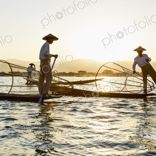 Three fisherman on Inle Lake, standing and using traditional rowing techniques,…