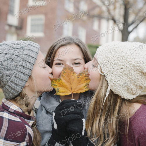 Three girls, one holding an autumn leaf by her face, two kissing her cheek. Three girls, one holding an autumn leaf by her face, two kissing her cheek.