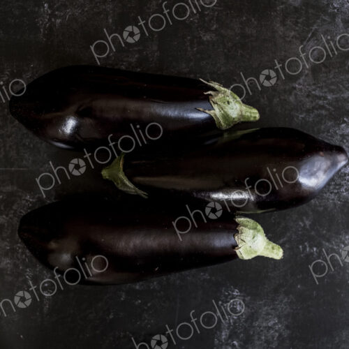 Three glossy purple aubergine egg plant vegetables on a grey slate background Three glossy purple aubergine egg plant vegetables on a grey slate background